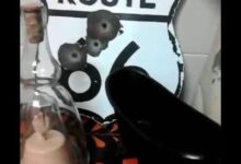 Cartel Route 66 con marcas de impacto, botella de cristal con vela y zapato de tacón negro en un montaje de feria — Feria Desembalaje de Antigüedades de Murcia