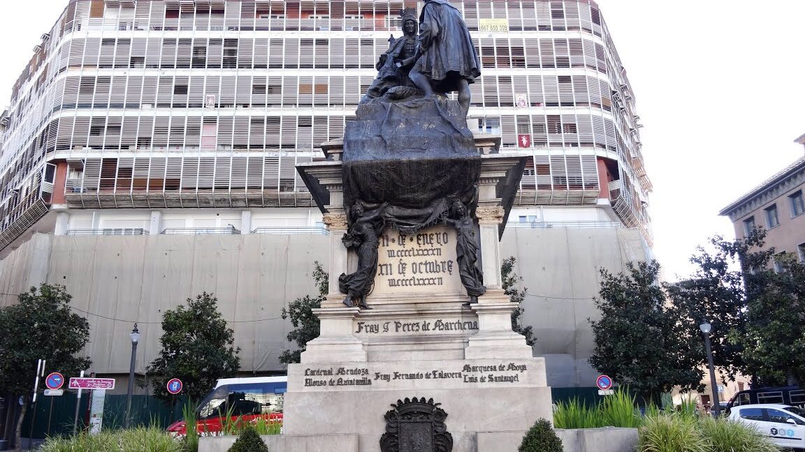 Monumento en plaza con edificios modernos, árboles y jardineras, imagen promocional para la Feria de Dulces Navidad de la Plaza Isabel II.