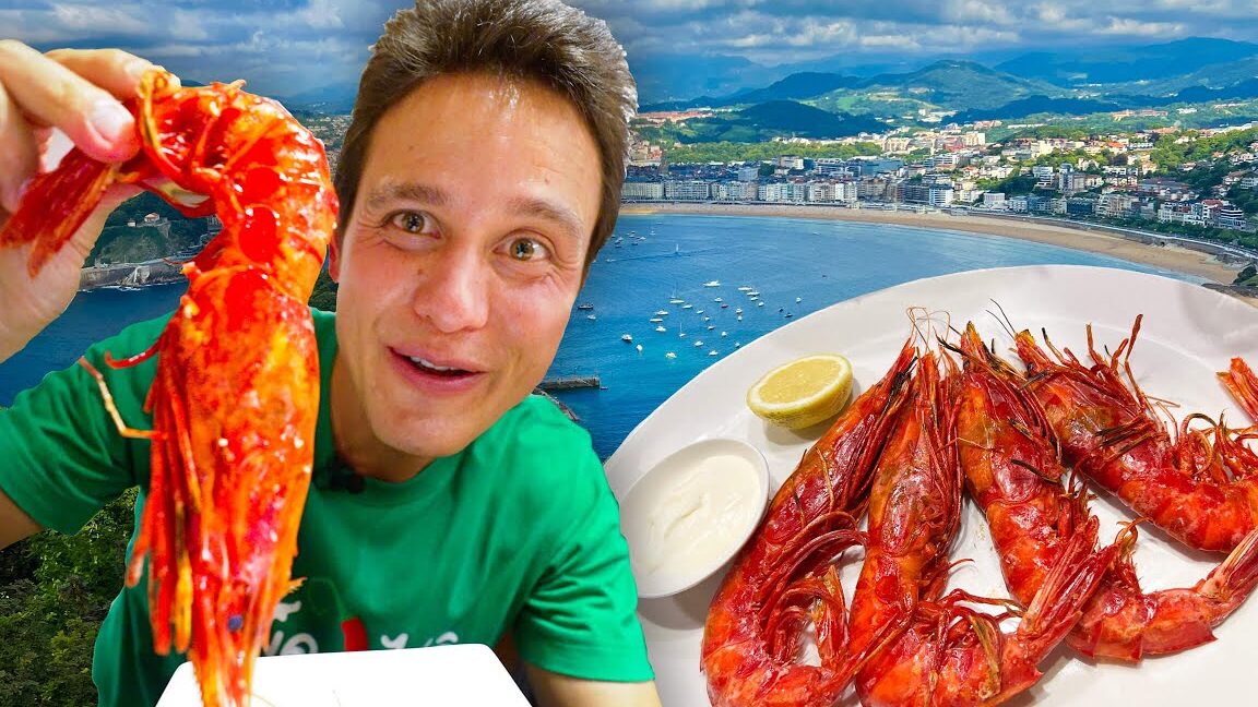 Persona sonriente mostrando una gamba grande junto a un plato de gambas y fondo de bahía en el Mercado Navideño de San Sebastián de los Reyes